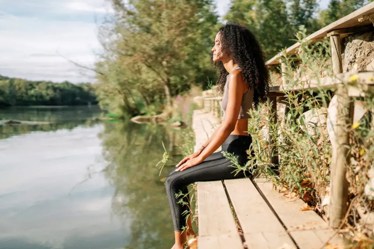 A woman sits by a tranquil lake, eyes closed, embodying how to be present in nature's embrace.
