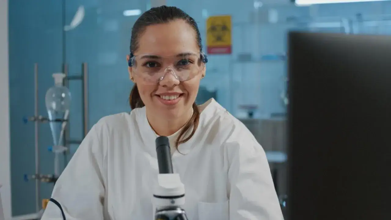 Uśmiechnięta kobieta w okularach ochronnych i białym fartuchu laboratoryjnym, przed mikroskopem. Zastanawia się, ile zarabia biolog.