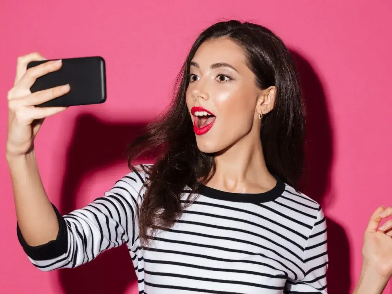 Kto to jest influencer? Młoda kobieta z czerwonymi ustami robi selfie telefonem na różowym tle.