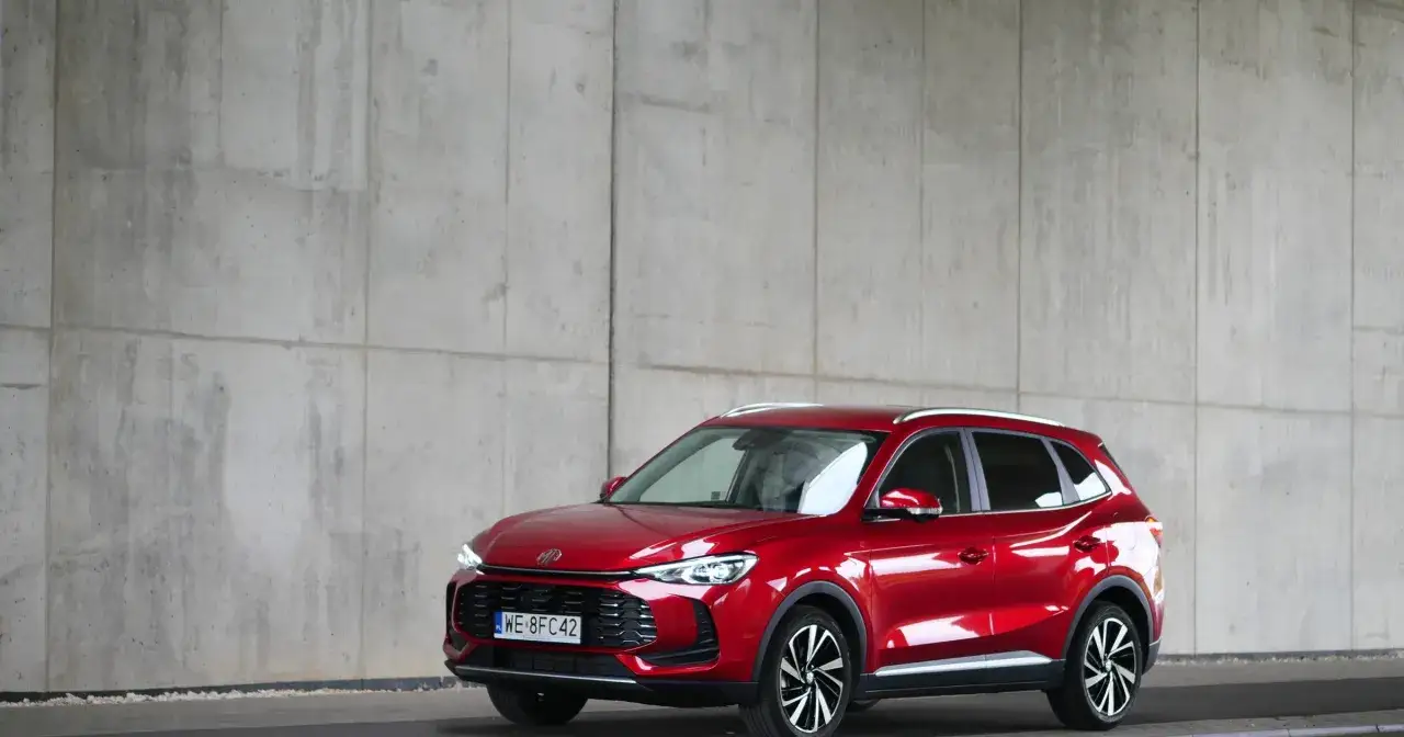 Błyszczący czerwony MG ZS Hybrid+ stoi przed betonową ścianą.