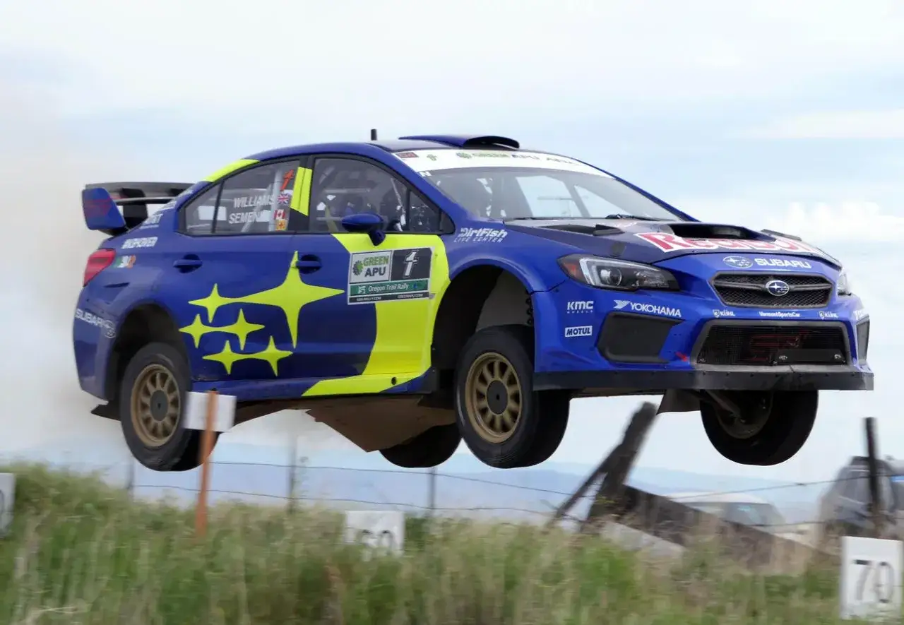 Blauer Subaru WRC mit gelben Sternen fliegt über eine Sprungschanze. Staub wirbelt auf.