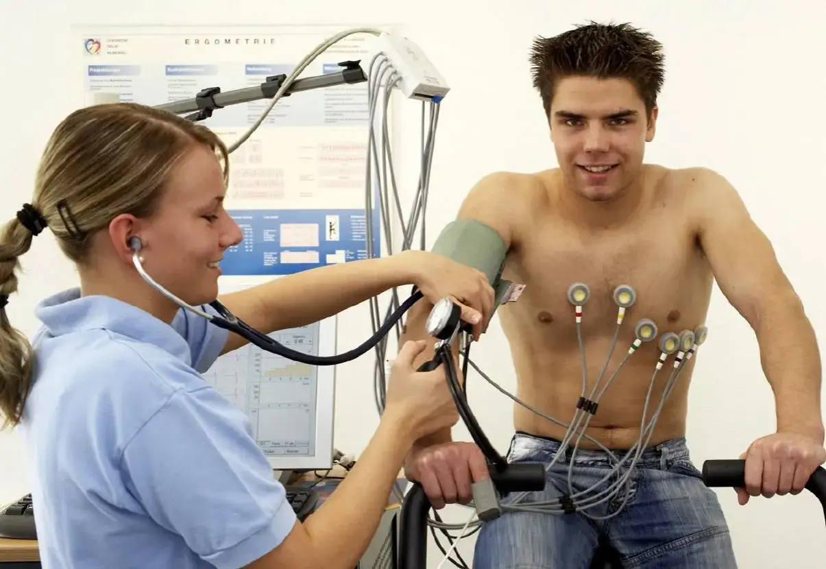 Mann unterzieht sich einem Belastungs-EKG auf einem Fahrradergometer, w&auml;hrend eine &Auml;rztin Blutdruck misst und EKG-Elektroden angebracht sind.