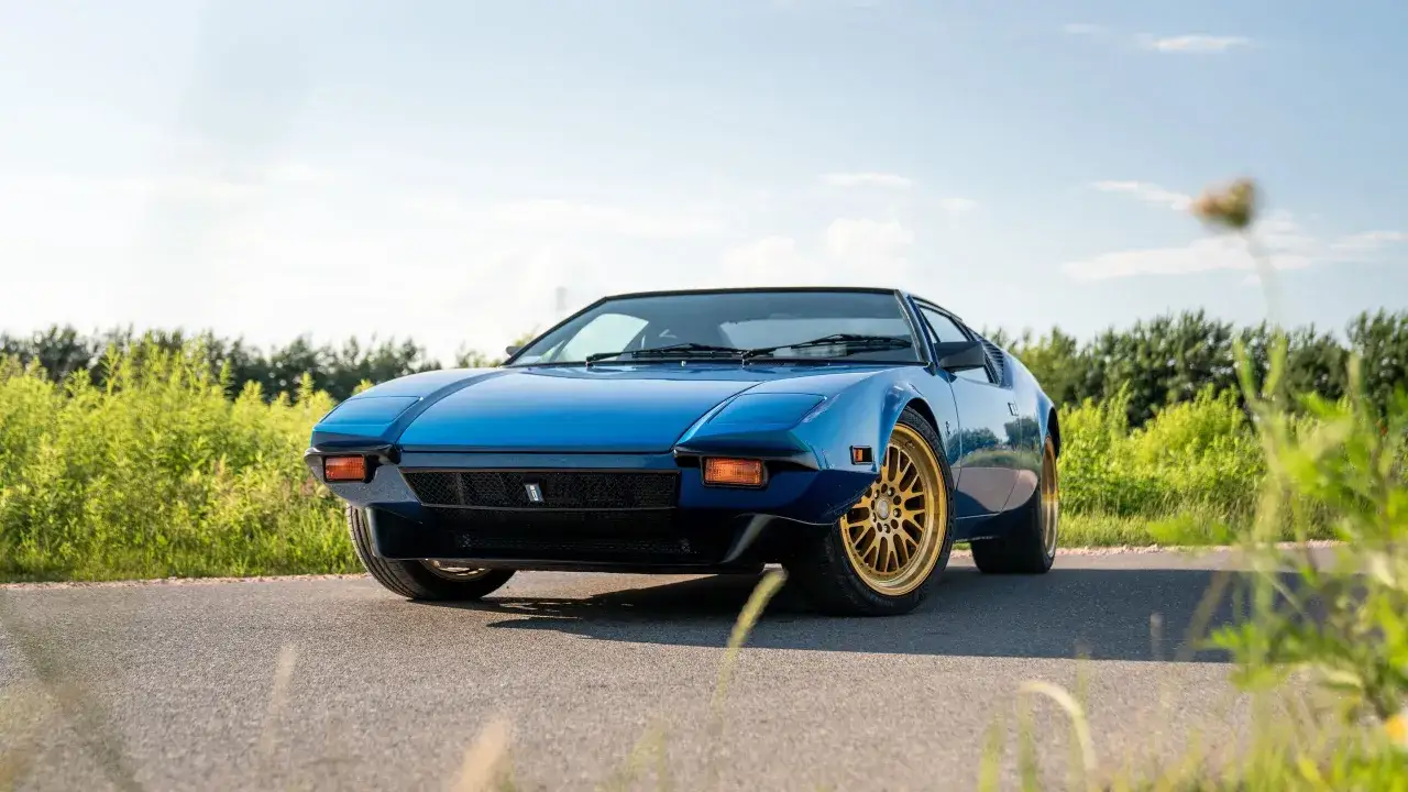 Niebieski De Tomaso Pantera z złotymi felgami na tle zieleni i błękitnego nieba.