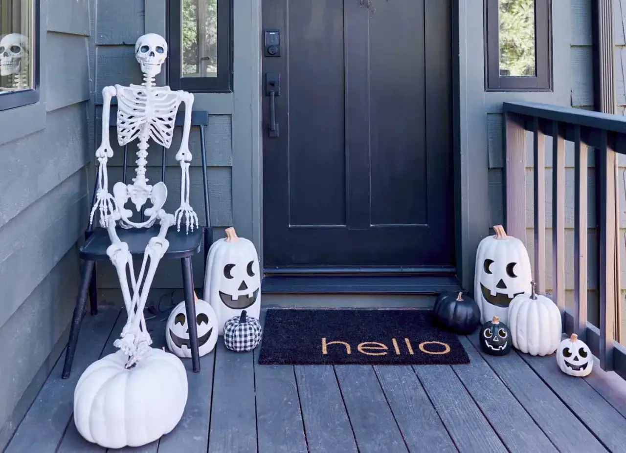 Un squelette s'assoit sur une chaise devant une porte, entouré de citrouilles blanches et noires. Une déco Halloween à faire soi-même qui accueille les visiteurs.