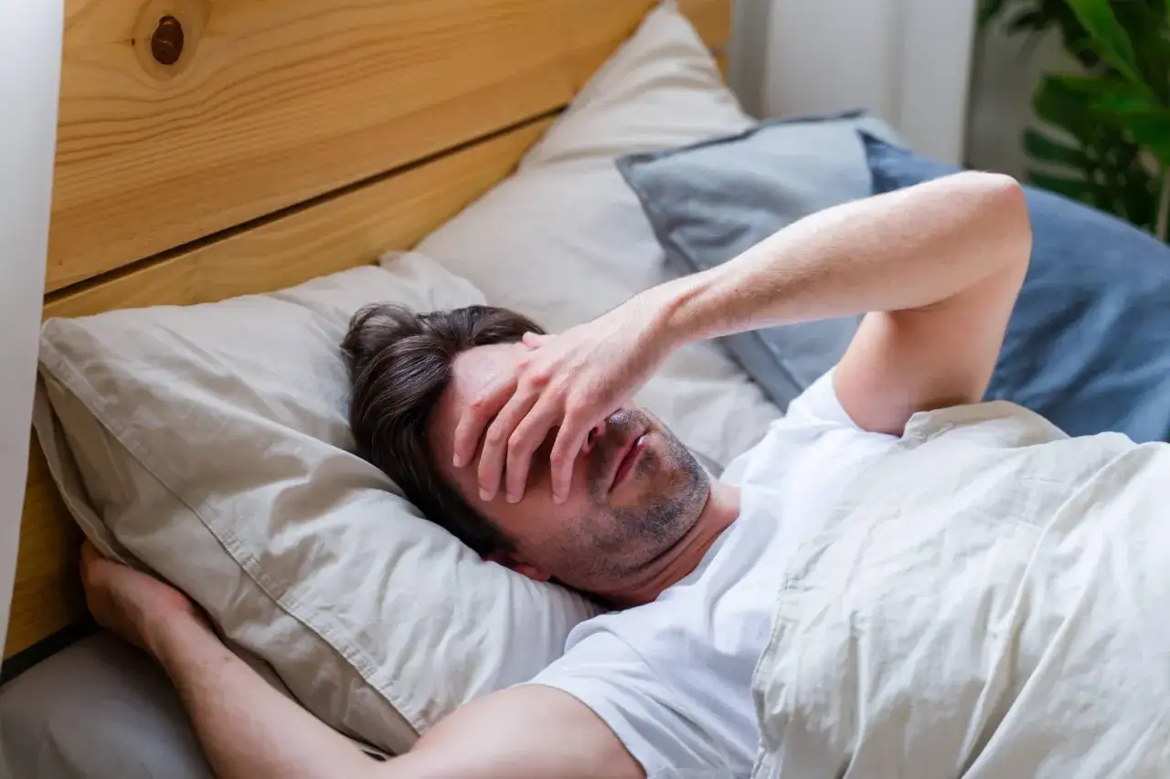 Mann liegt im Bett, hält sich die Hand vor die Augen. Er wirkt müde, als ob ihm nur sechs Stunden Schlaf nicht reichen.