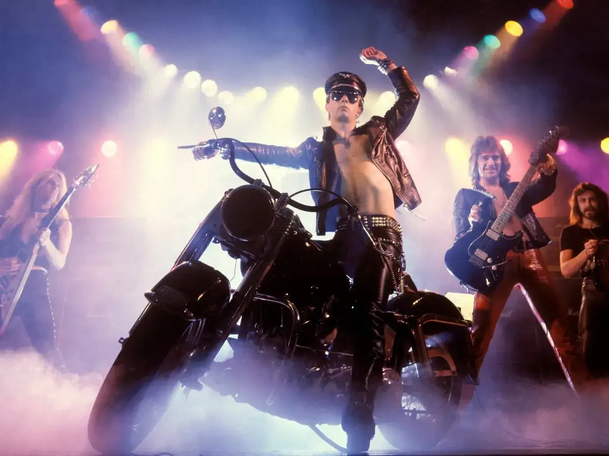 Judas Priest na scenie z motocyklem, grający swoje kultowe utwory. Wokalista w skórzanej kurtce i czapce, gitarzyści w rockowych strojach.