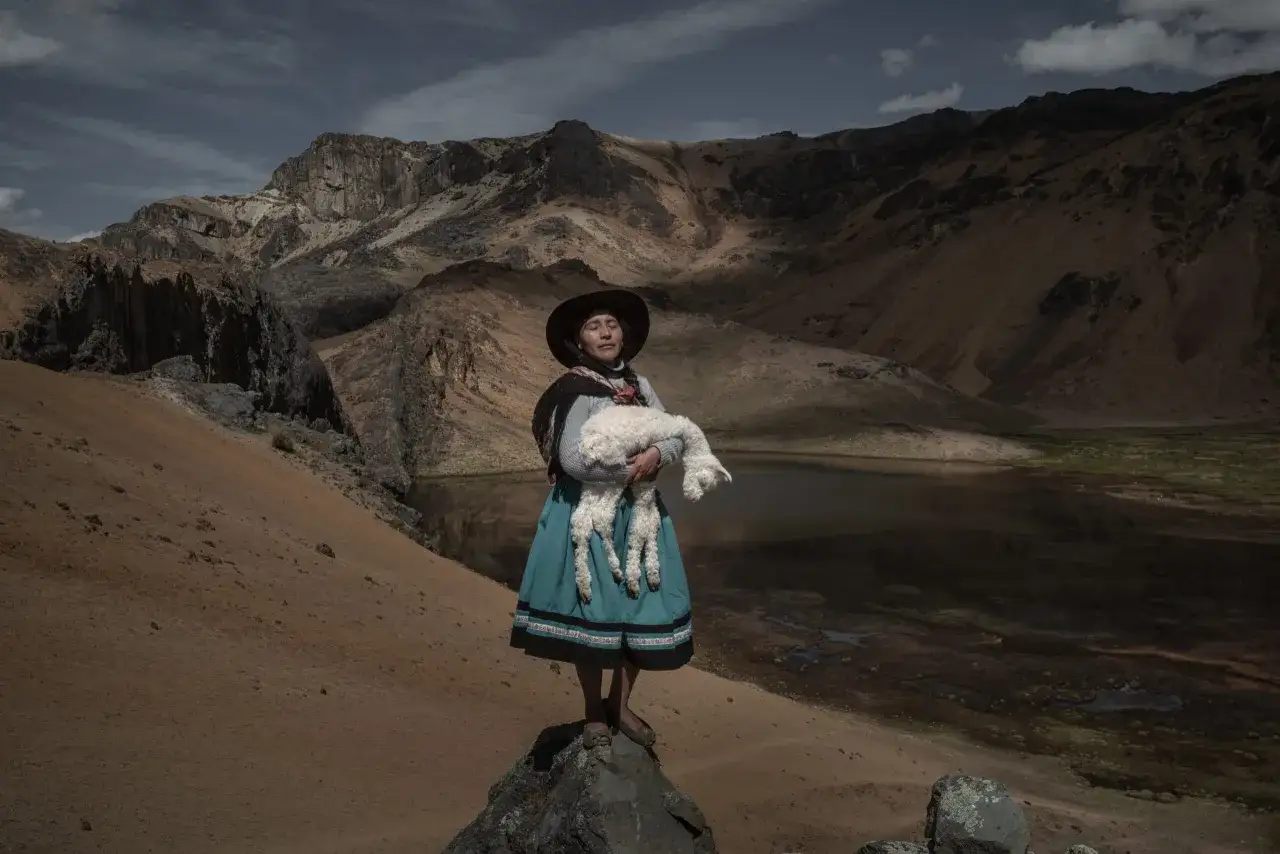 Donna con agnello in braccio, in piedi su una roccia, con montagne e lago sullo sfondo. Alessandro Cinque cattura la serenit&agrave;.