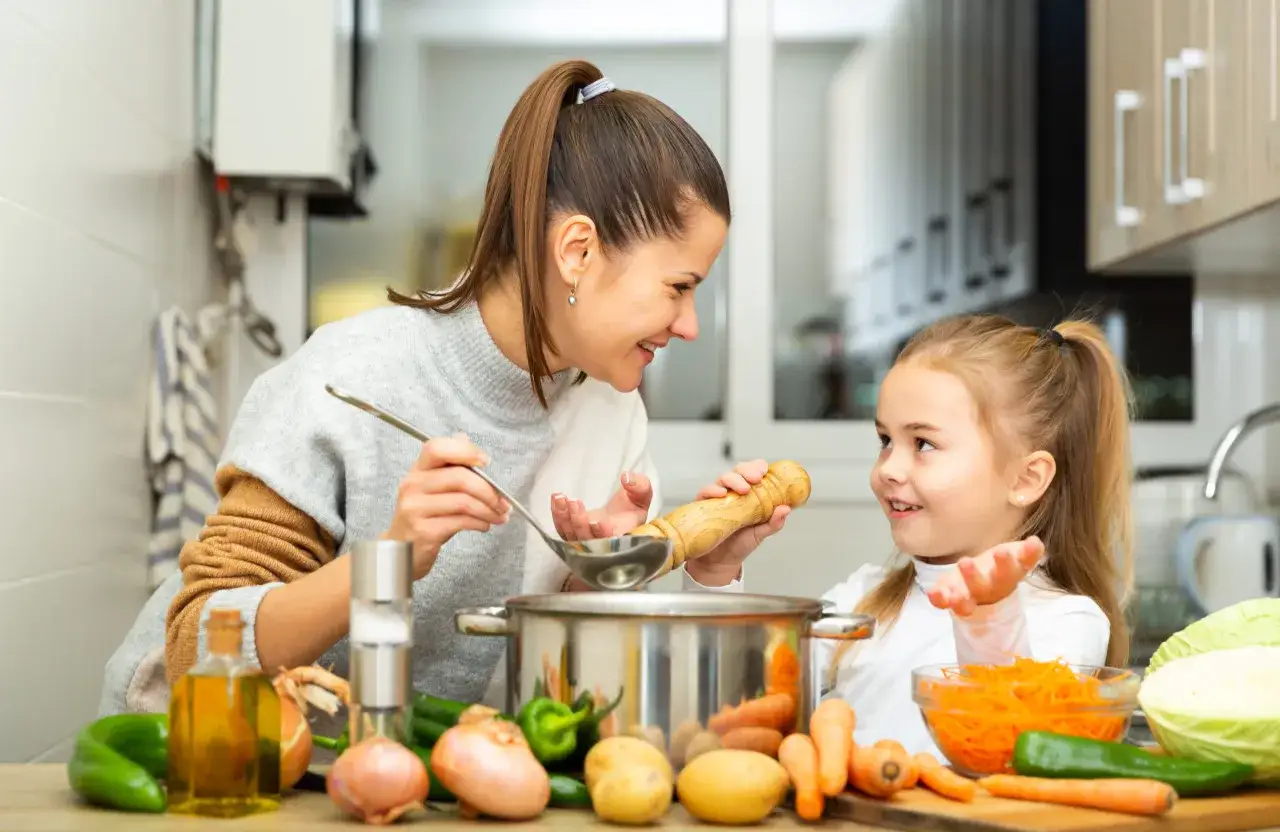 Mama i córka gotują pyszne zupy dla dzieci. Na stole warzywa: marchewka, ziemniaki, cebula, papryka i kapusta.