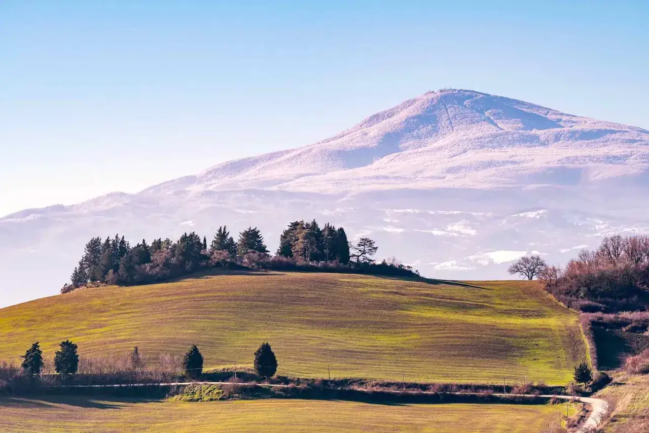 Paesaggio toscano con campi verdi e il Monte Amiata innevato in lontananza. Immagina la **funivia Monte Amiata** che sale verso la cima.