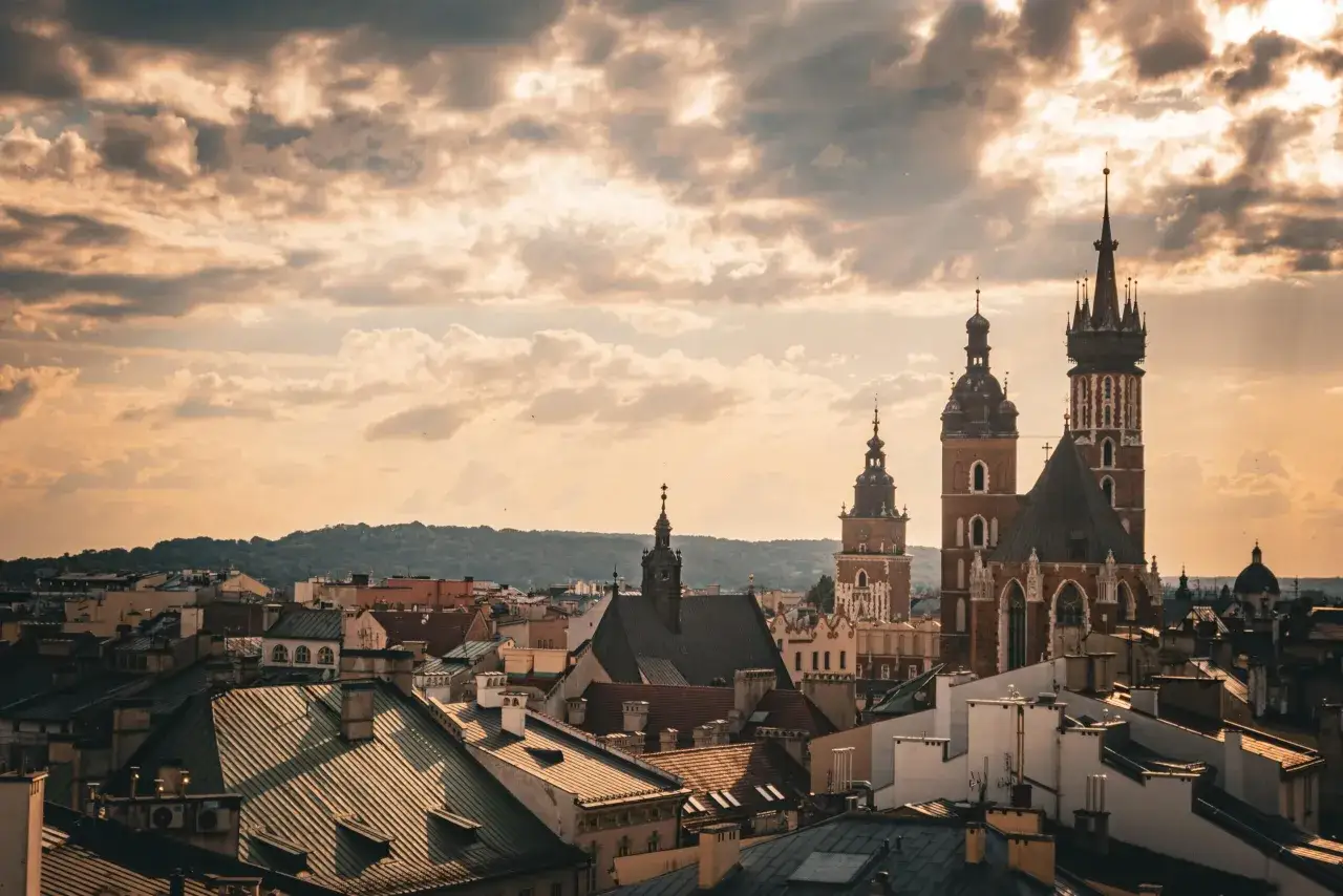 Widok na krakowskie dachy i wieże kościołów pod burzowym niebem. W oddali widać wzgórza.