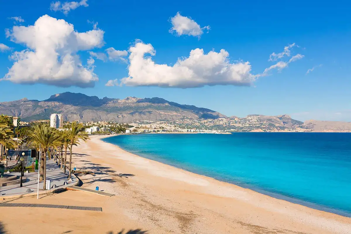 Playa de arena dorada, palmeras y mar turquesa bajo un cielo azul con nubes. As&iacute; es cual es la costa blanca, un para&iacute;so mediterr&aacute;neo.