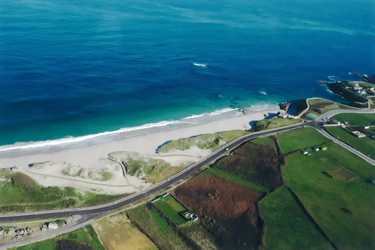 Vista aérea de la **playa llas** con arena blanca, mar azul y campos verdes. Una carretera serpentea junto a la costa.