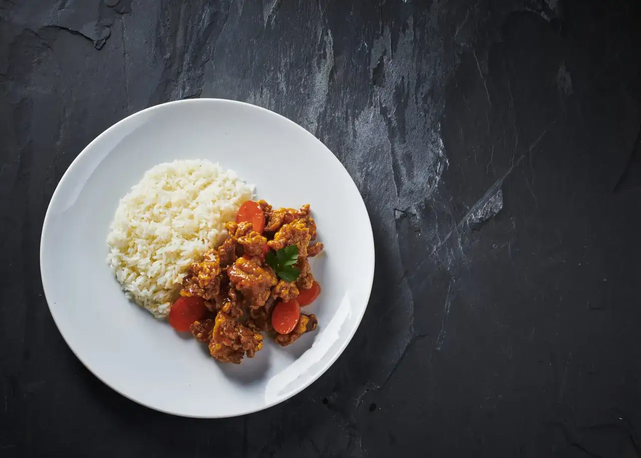 En tallrik med ris och krispig orange chicken, garnerad med persilja och morotsbitar. En l&auml;cker m&aring;ltid som ser ut att vara redo att avnjutas.