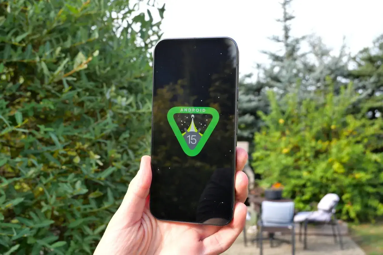 Dłoń trzyma smartfon z logo **Android 15** na ekranie, na tle zieleni.
