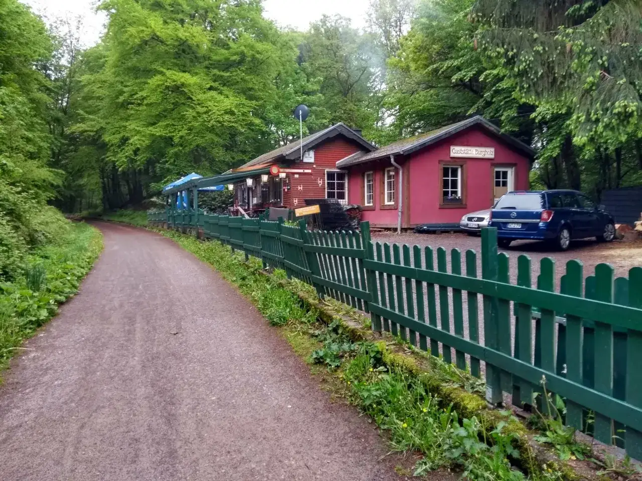 Ein Weg f&uuml;hrt zum Gasthof Burgholz, umgeben von gr&uuml;nen B&auml;umen. Ein blauer Kombi parkt davor.