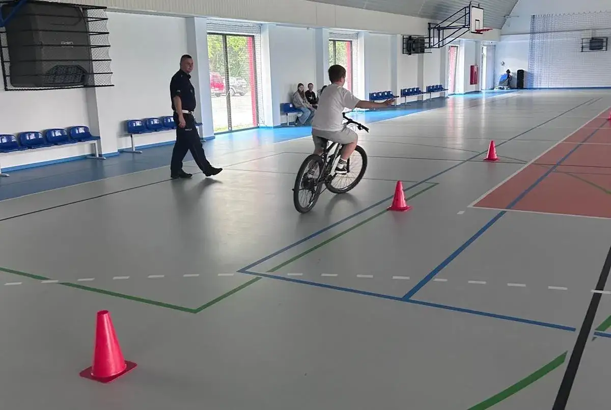 Chłopiec na rowerze pokonuje tor przeszkód na hali sportowej, przygotowując się do egzaminu na kartę rowerową. Obok stoi policjant.