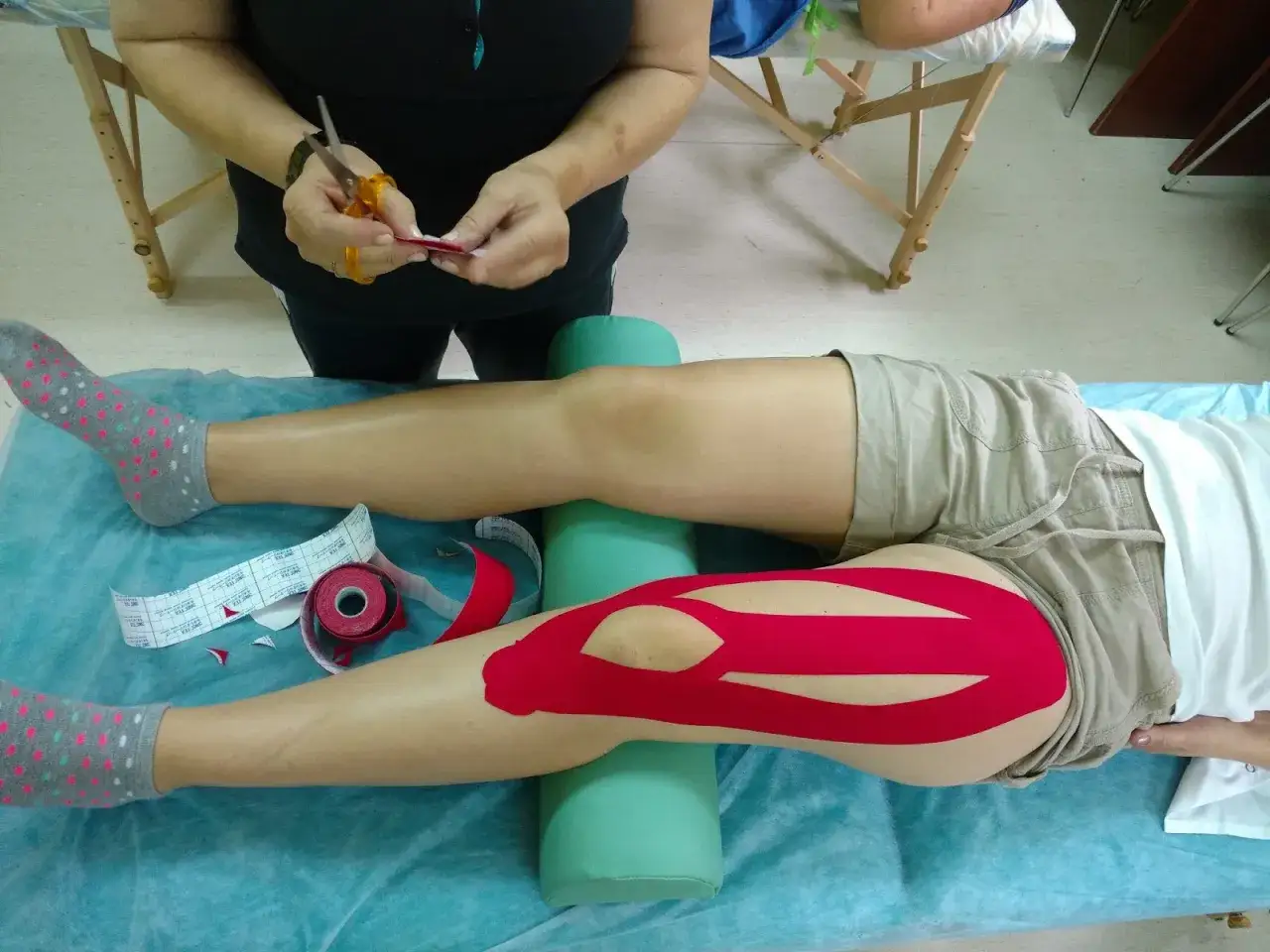 Taping mięśnia czworogłowego uda czerwonymi plastrami. Terapeuta przygotowuje taśmę do aplikacji.