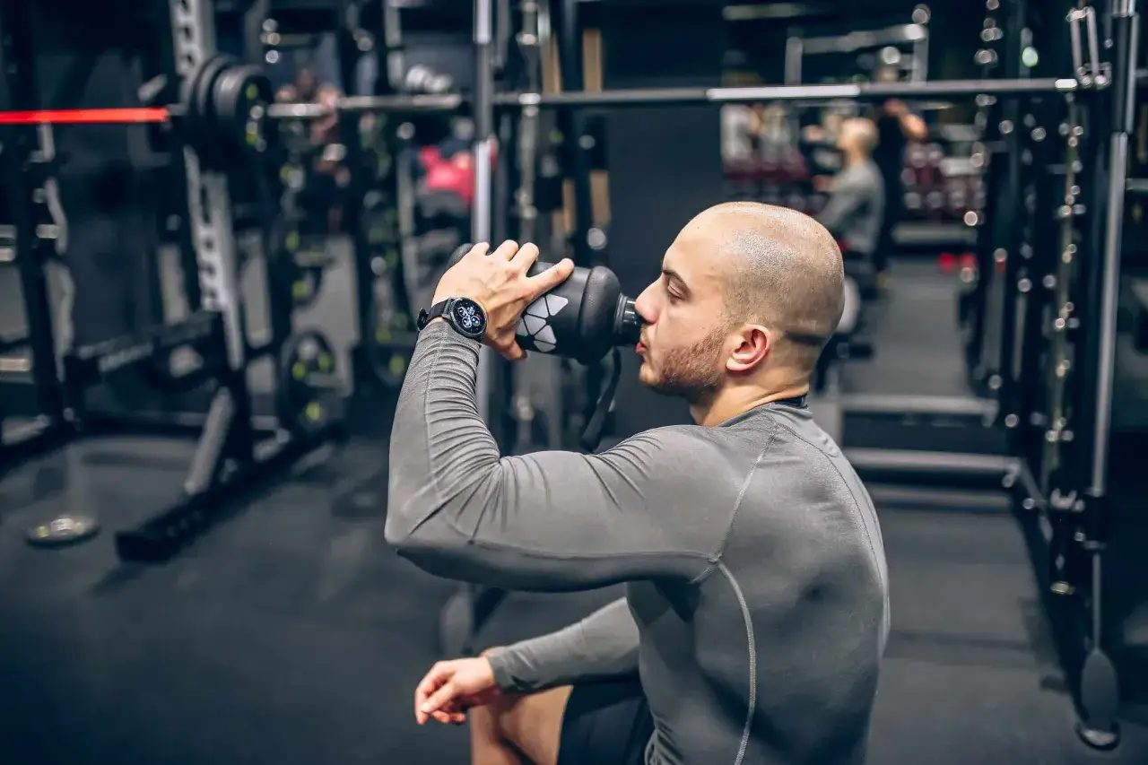 Uomo con orologio sportivo beve da una borraccia in palestra, pensando a proteine prima o dopo allenamento per recuperare.