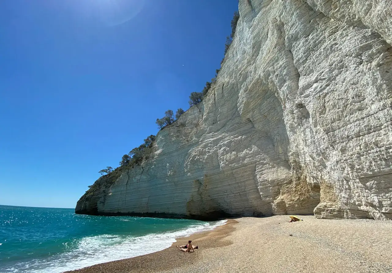Una persona si rilassa sulla spiaggia di ciottoli di una cala rosa a Mattinata, con imponenti scogliere bianche e mare turchese.
