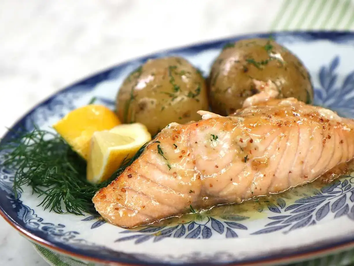 En lyxig hel lax i ugn, perfekt tillagad med citron och dill. Serveras med små, örtkryddade potatisar på en vacker blåvit tallrik.