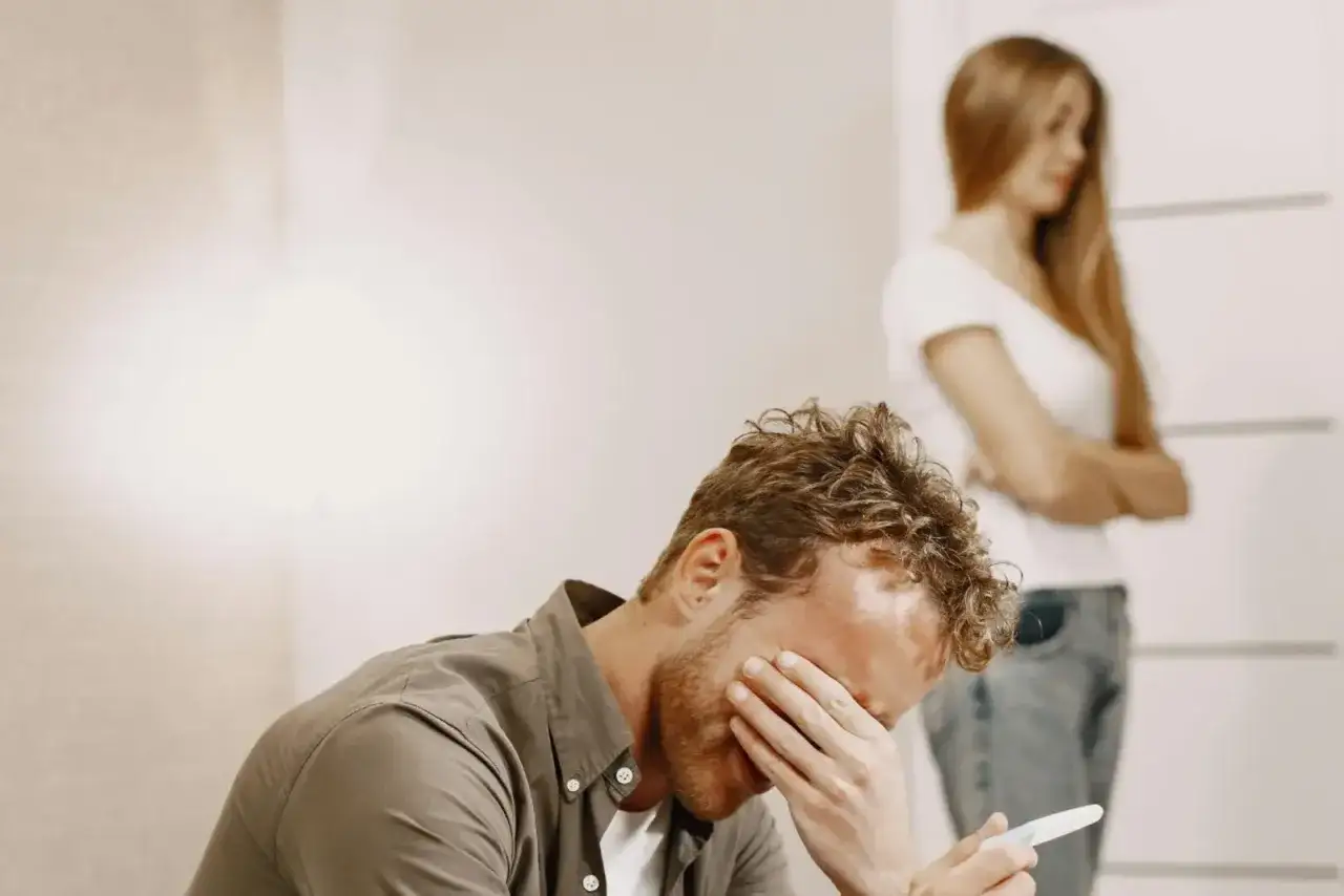 Uomo disperato con test di gravidanza in mano, donna con braccia conserte sullo sfondo. Forse si pentirà di credere di non amare più.