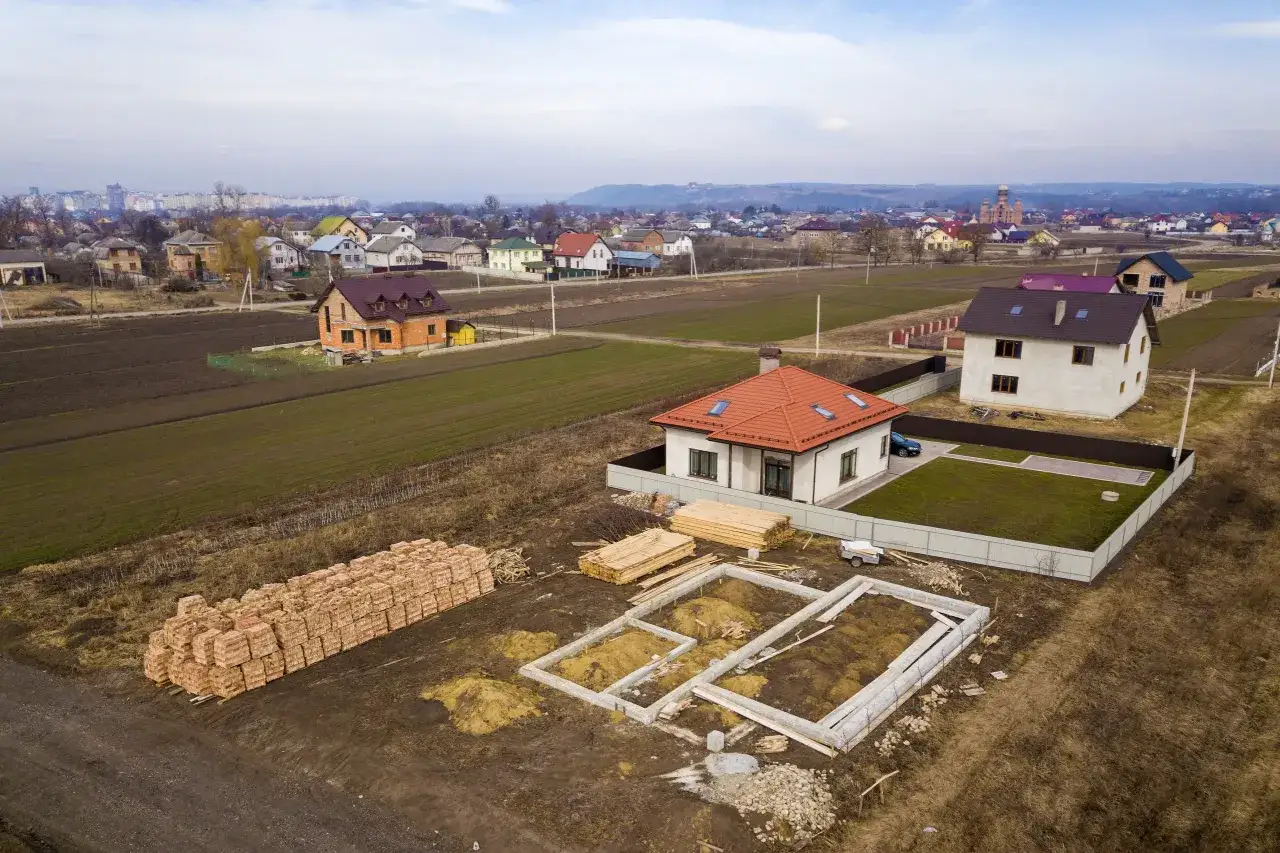 Pusty plac budowy z fundamentami, stosem cegieł i drewnem. W tle domy i miasto. To dobry początek, by dowiedzieć się, co zrobić po zakupie działki budowlanej.