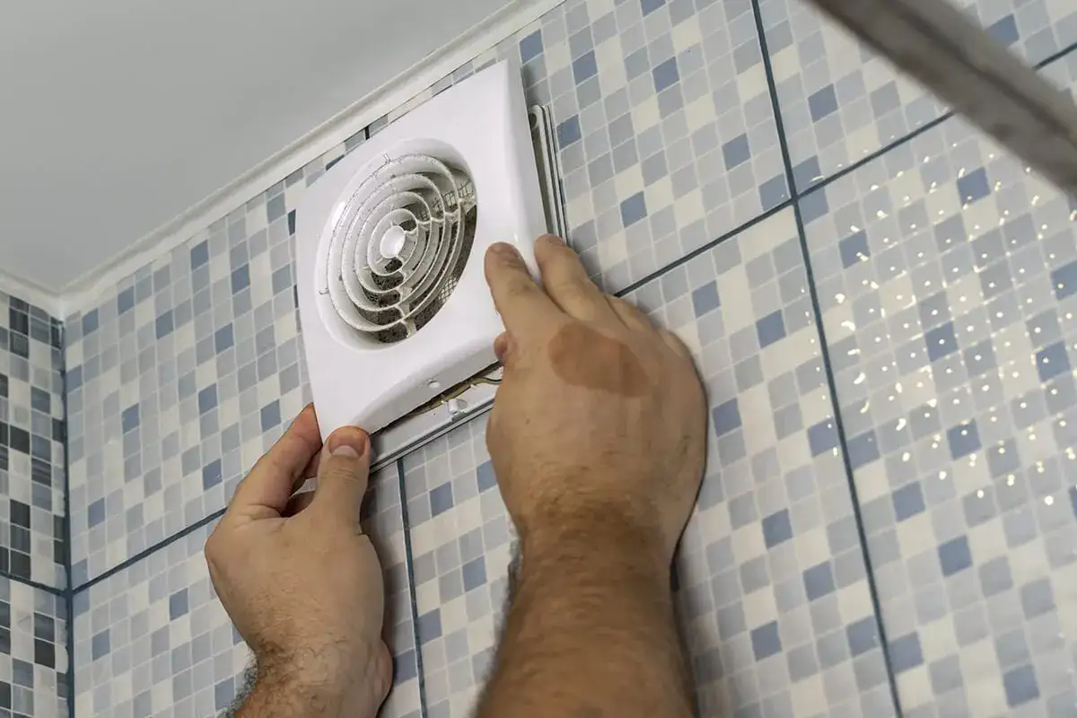 Installation d'un VMC salle de bain norme. Un homme ajuste un extracteur d'air blanc sur un mur carrelé bleu et blanc.