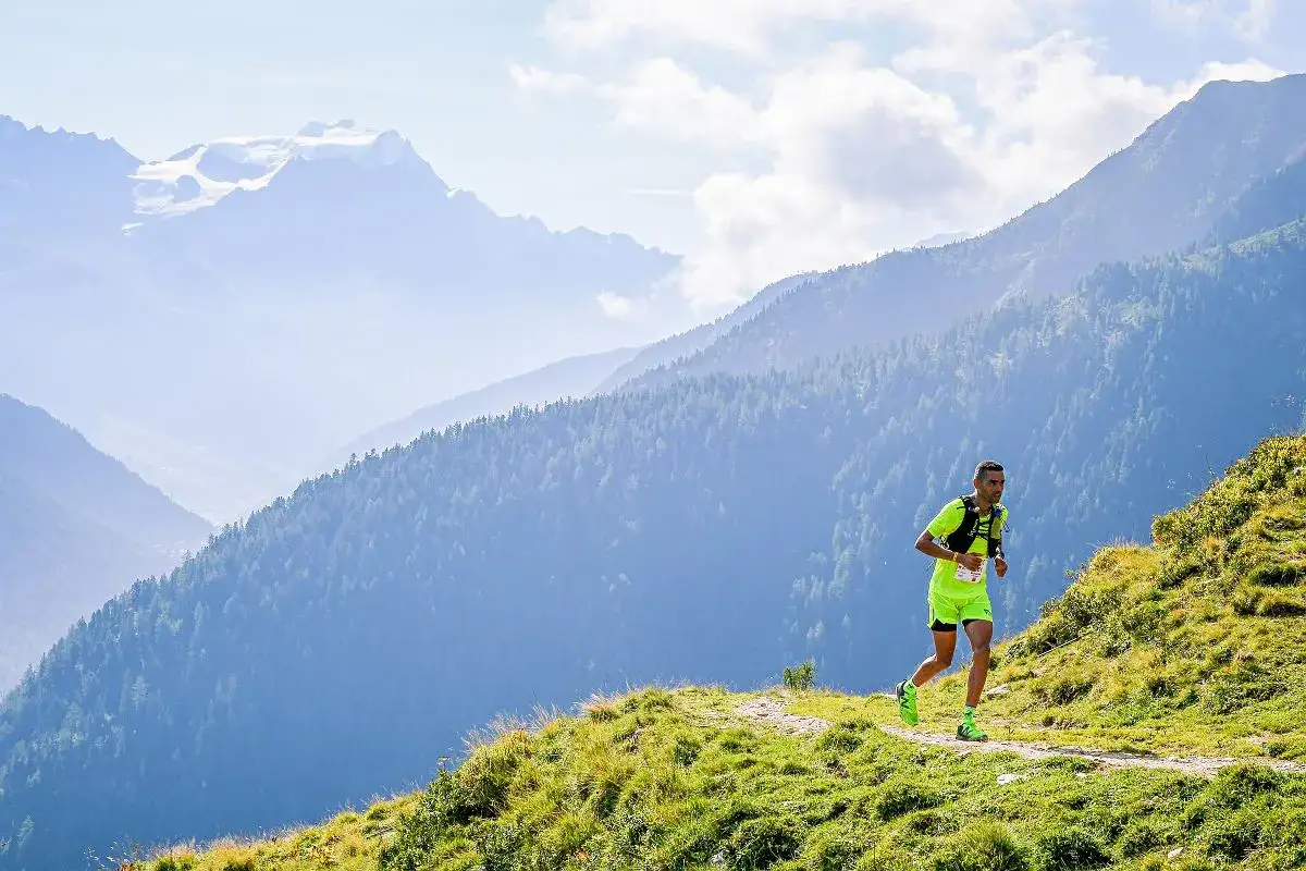 Biegacz w jaskrawej koszulce pokonuje trasę ultra-trail du mont-blanc, otoczony majestatycznymi, zalesionymi górami i ośnieżonymi szczytami w tle.