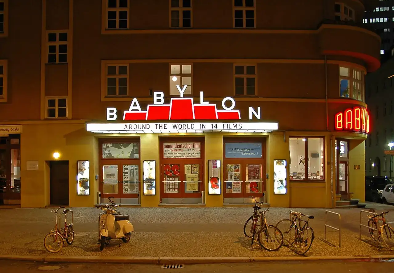 Das Babylon Kino Berlin bei Nacht, mit leuchtender Neonreklame und Filmplakaten. Fahrr&auml;der und ein Roller stehen davor.