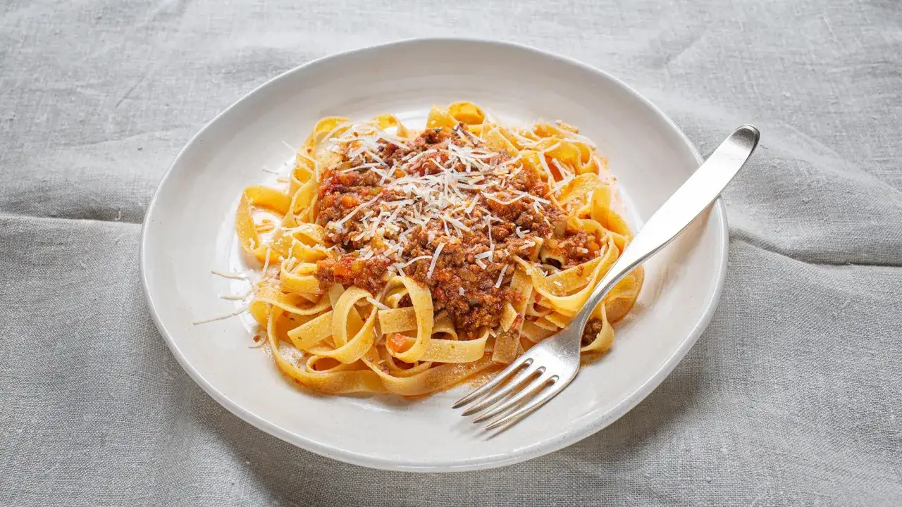 Fettuccine alla bolognese mit geriebenem Parmesan auf einem Teller, daneben eine Gabel.