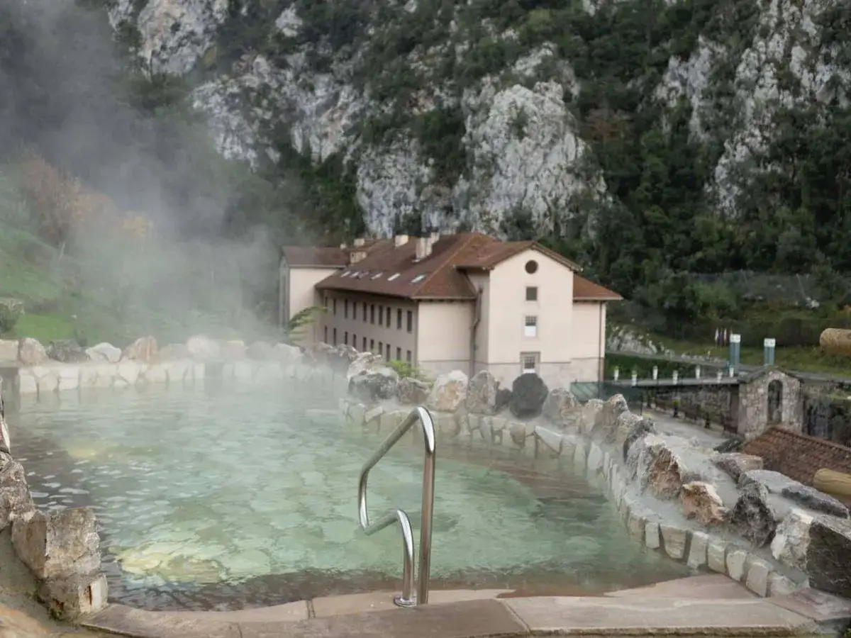 Piscina de aguas termales en España, rodeada de rocas y con vapor ascendiendo. Al fondo, un edificio y montañas verdes.