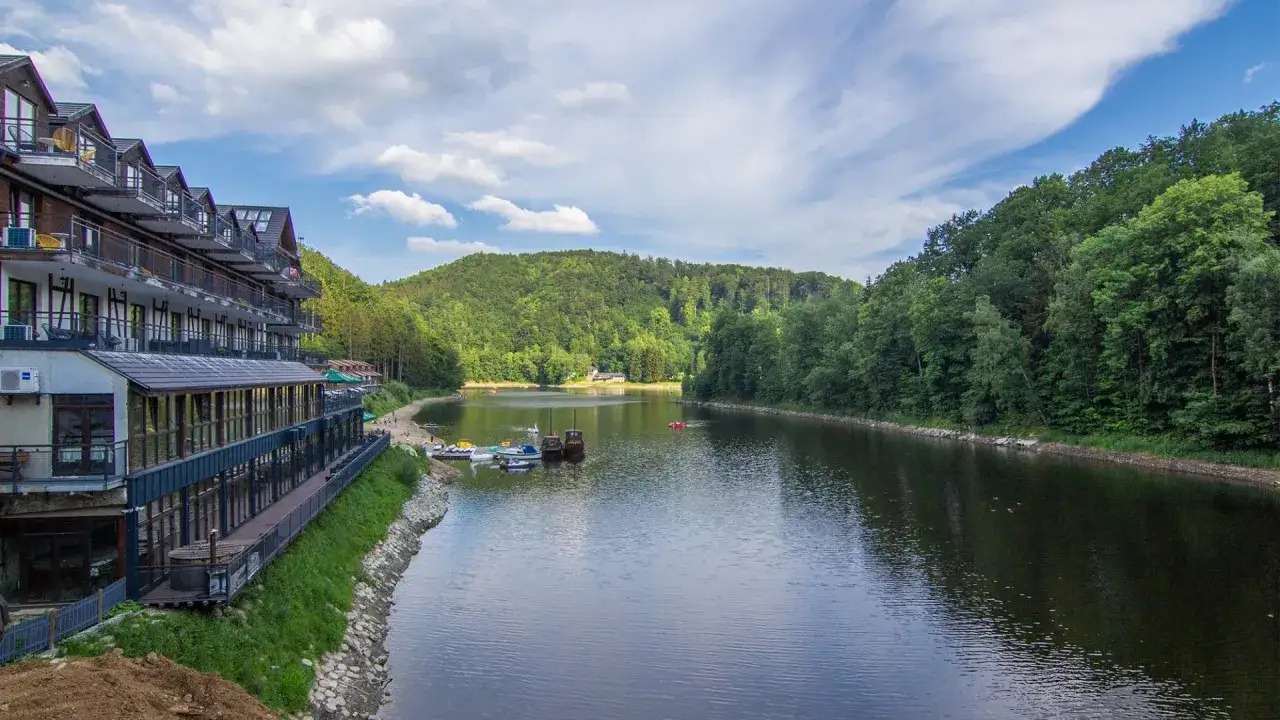Hotel nad jeziorem w Zagórzu Śląskim. Woda odbija zielone wzgórza i chmury. Na wodzie kilka łódek.