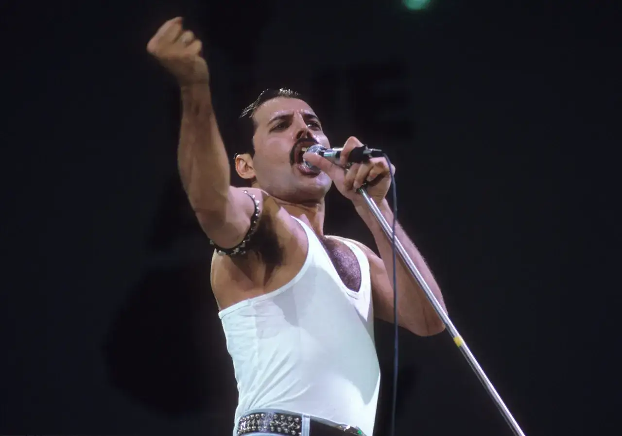 Freddie Mercury w białej podkoszulce, z uniesioną pięścią, śpiewa na jednym z niezapomnianych **Queen koncerty**.