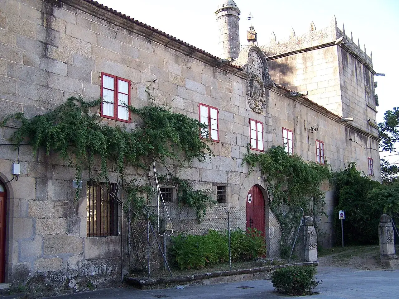 El pazo Vista Alegre, con sus muros de piedra y ventanas rojas, se cubre de vegetación. Un portón rojo da la bienvenida a este histórico lugar.