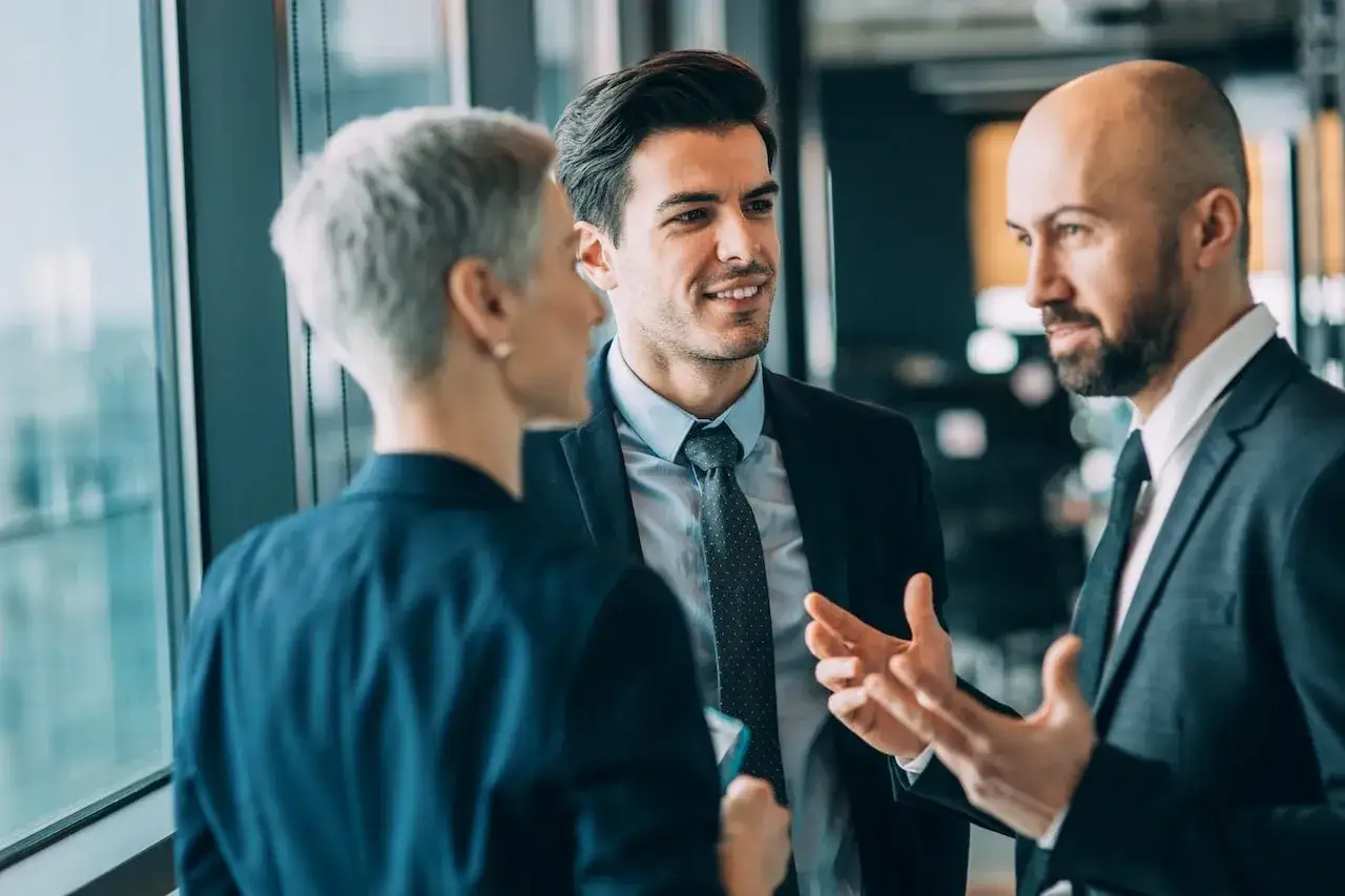 Three professionals engage in influential communication, discussing ideas near a large window.