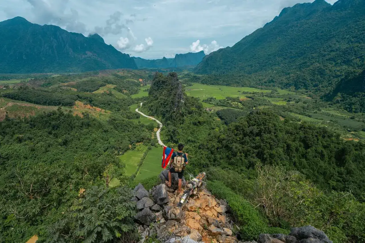 Ein Backpacker mit Rucksack und Laos-Flagge steht auf einem Felsen und blickt auf ein grünes Tal.