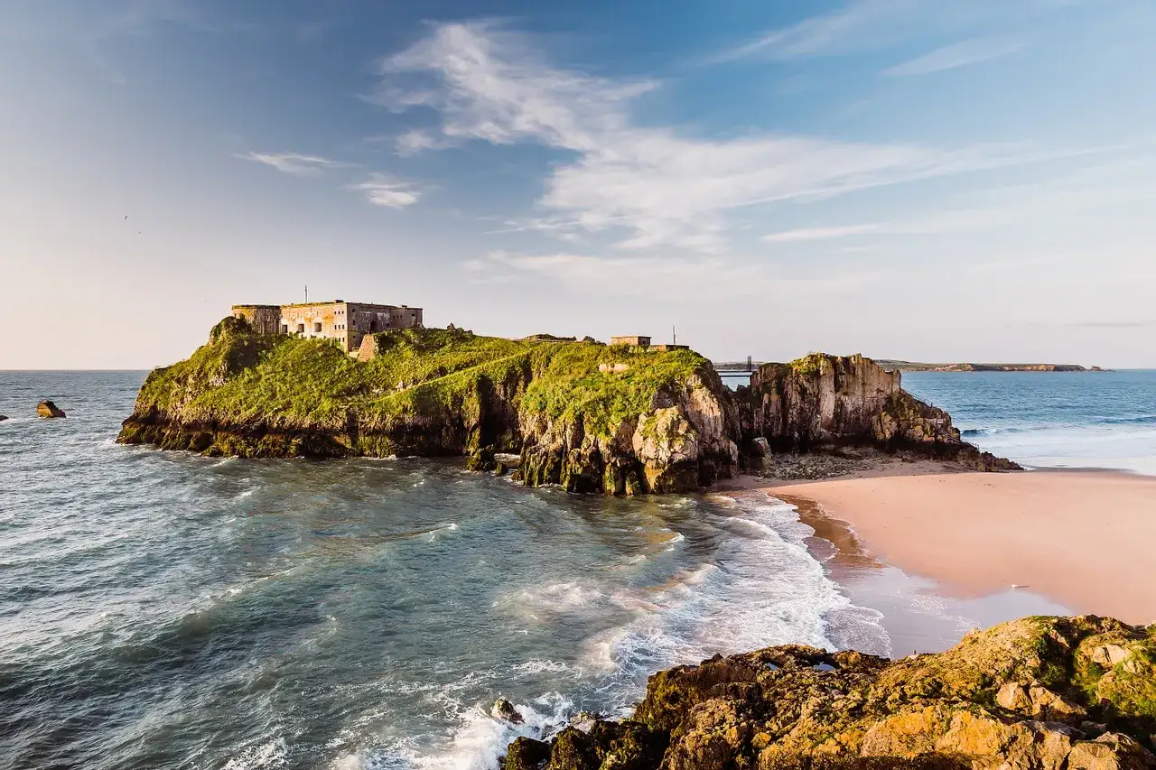 Fortification sur une île verdoyante, bordée par une plage de sable rose et une mer agitée. Un lieu idéal pour un séjour au Pays de Galles.