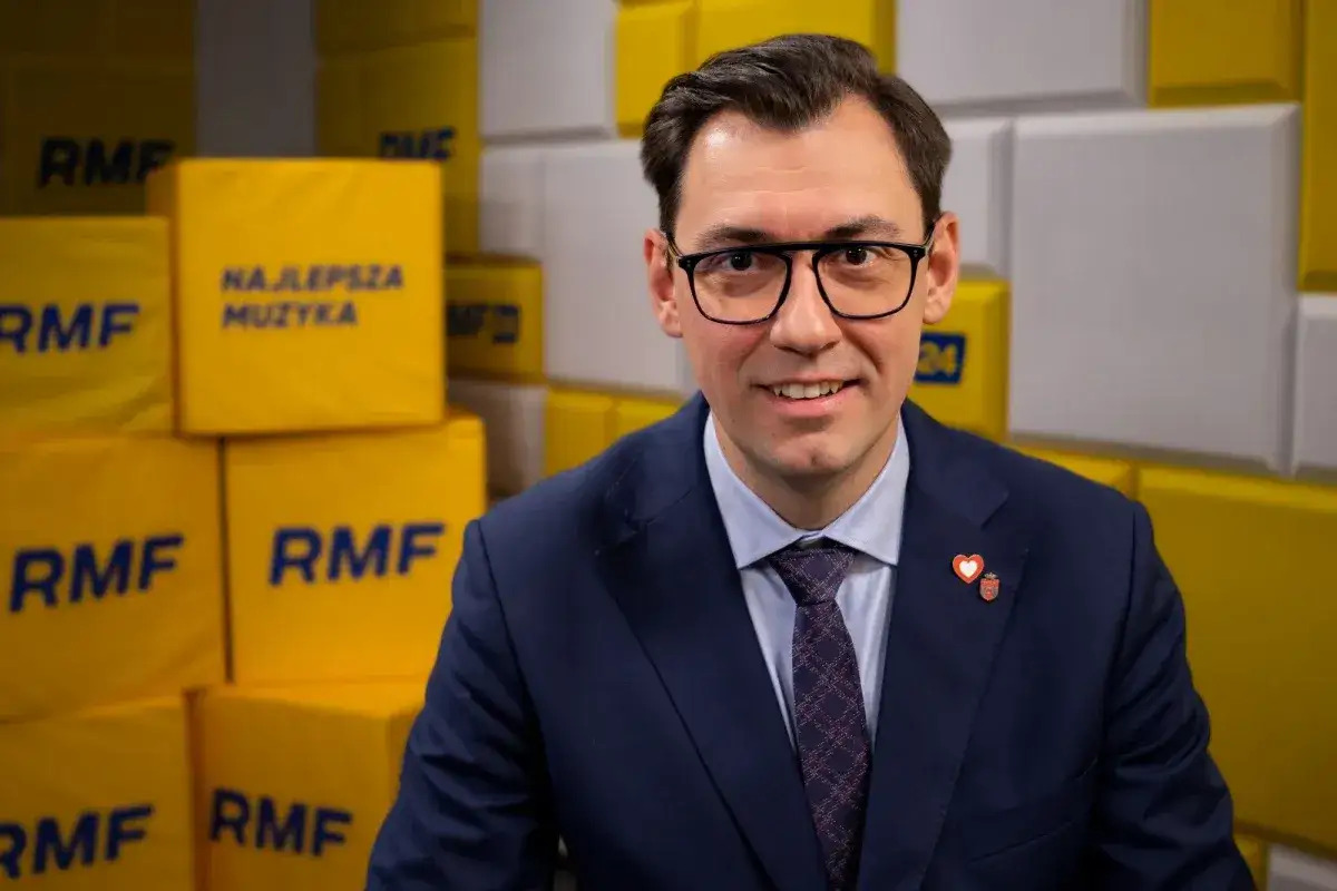 Konrad Frysztak, z wykształceniem, w garniturze, na tle żółtych kostek z logo RMF.