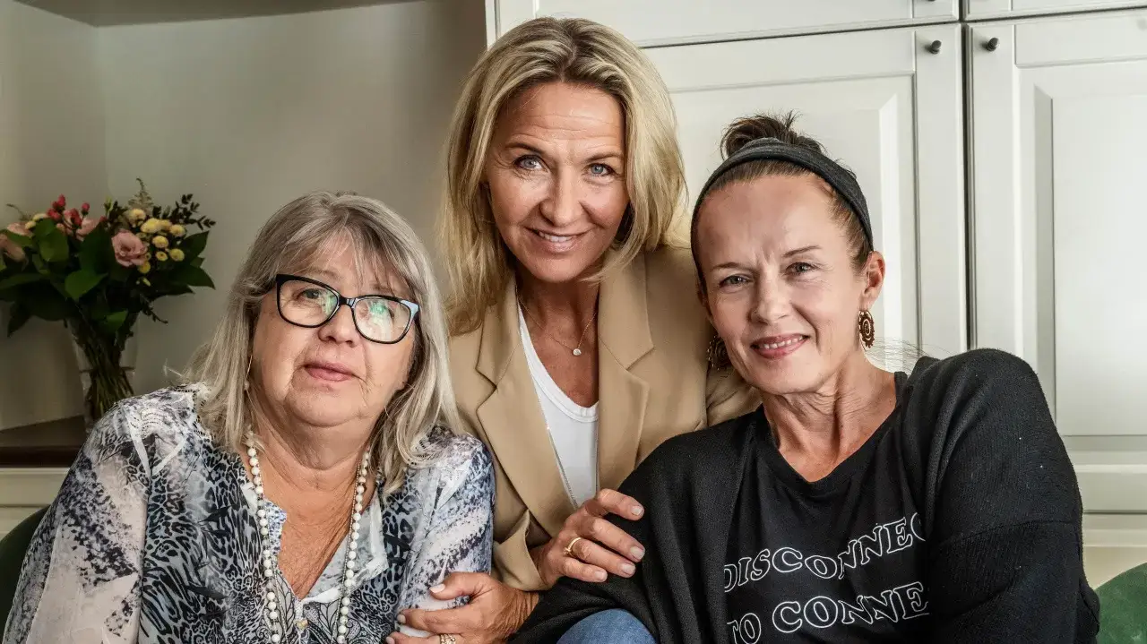 Tre kvinnor, kanske lill babs barn, ler mot kameran. En kvinna i mitten bär en beige kavaj, den vänstra har glasögon och den högra en svart tröja.