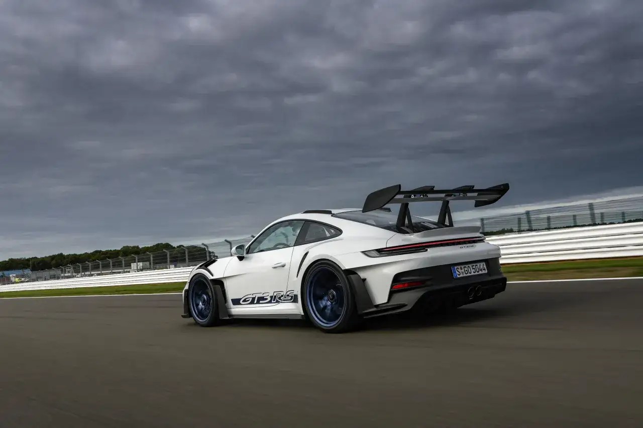 Białe Porsche 911 GT3 RS z niebieskimi felgami i naklejkami GT3 RS pędzi po torze wyścigowym pod pochmurnym niebem.
