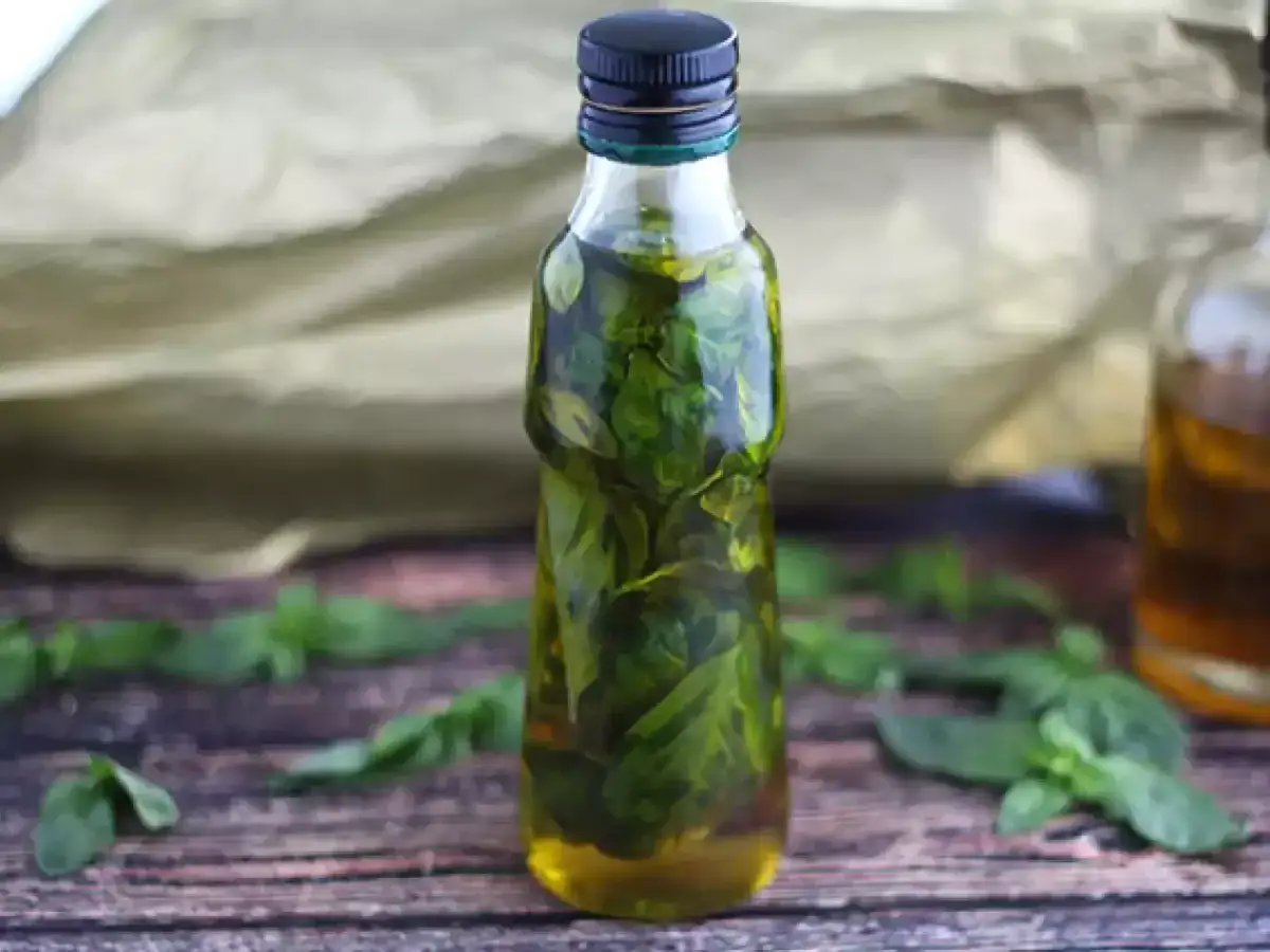 Bottiglia di vetro piena di olio al basilico fresco, con foglie sparse sul tavolo di legno.