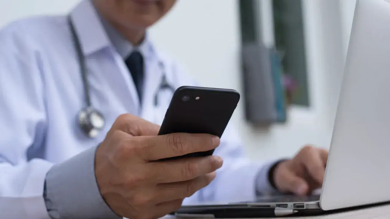 Lekarz w białym kitlu z telefonem w dłoni, obok laptop. Szybka konsultacja l4 przez telefon.