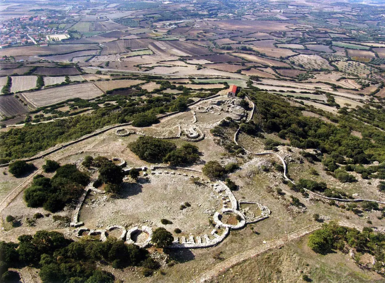 Vista aerea del **santuario nuragico di Santa Vittoria**, con le sue antiche strutture in pietra e un piccolo edificio rosso in cima alla collina.