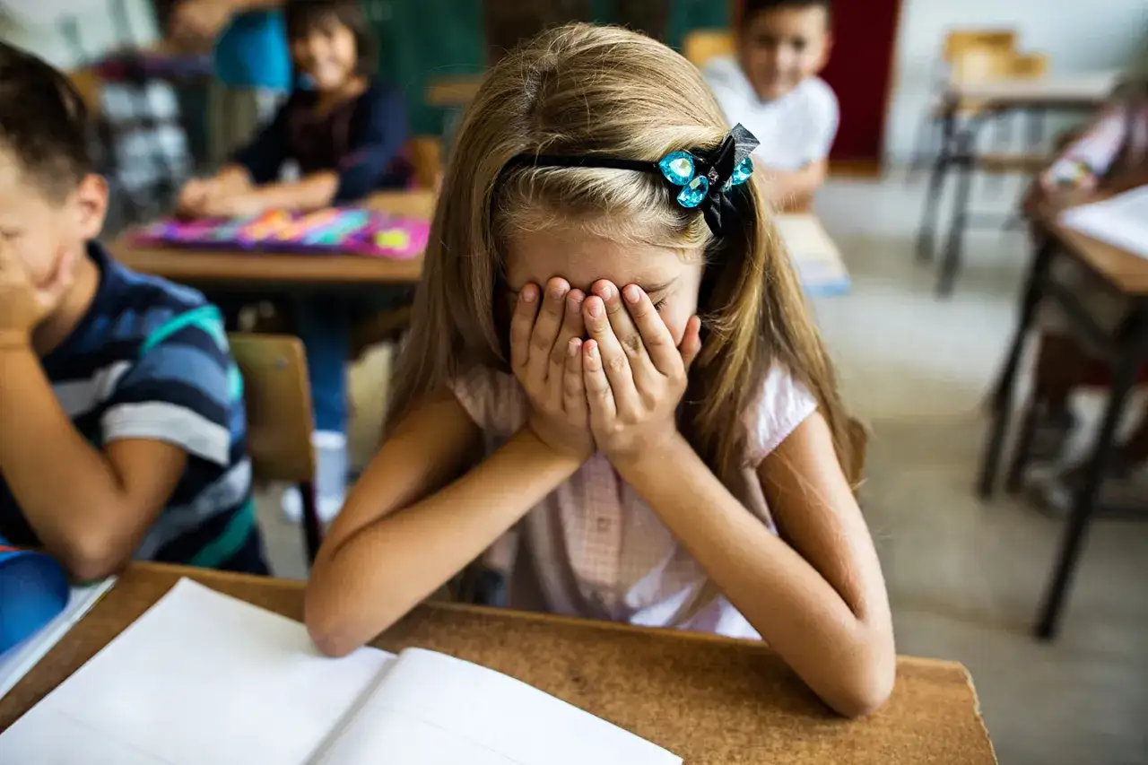 Ein M&auml;dchen versteckt ihr Gesicht in den H&auml;nden, Schulangst was tun? Andere Kinder sitzen im Hintergrund.