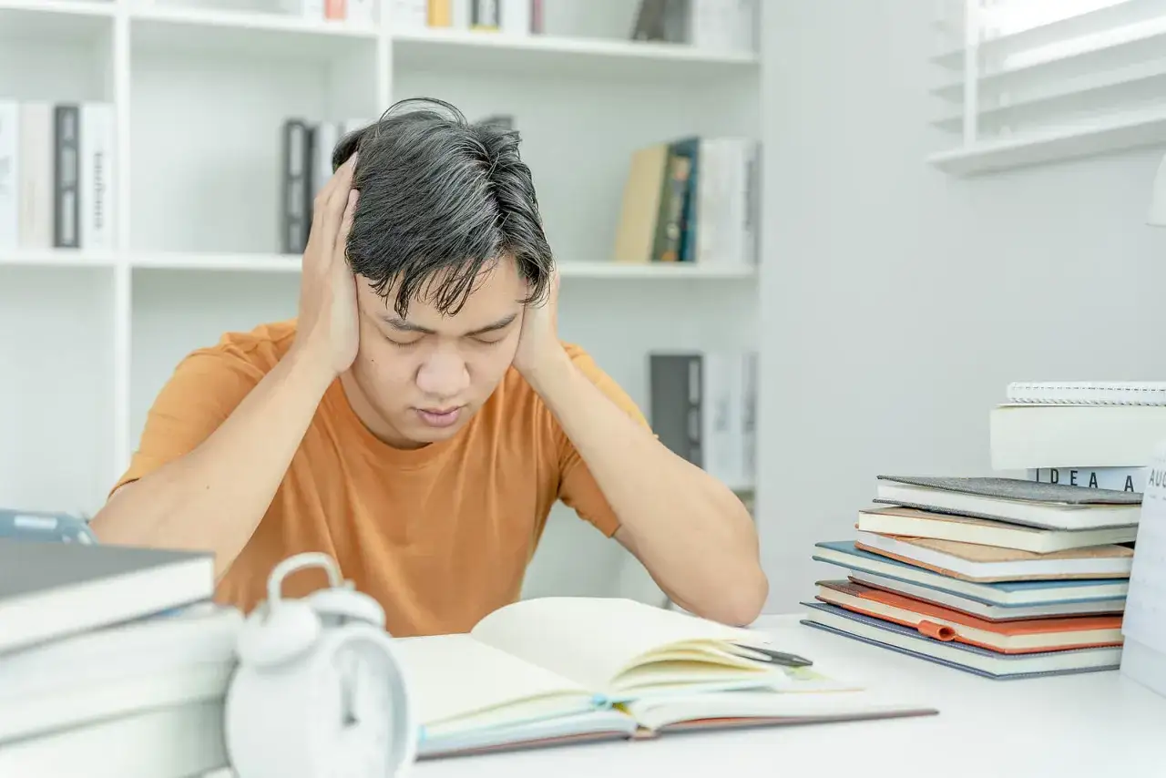 Ein junger Mann mit Händen an den Schläfen, der über einem offenen Buch sitzt. Er leidet offensichtlich unter Prüfungsangst.