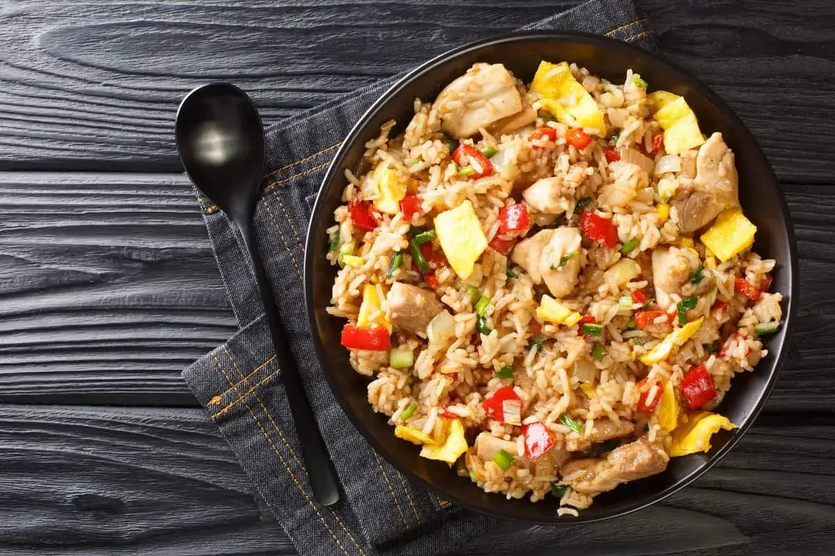 Un plato de arroz chaufa con trozos de pollo, huevo revuelto, pimientos rojos y cebolla verde, servido en un tazón negro junto a una cuchara.