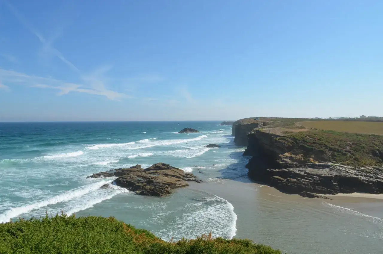 Costa salvaje con acantilados y mar turquesa en la playa de Esteiro, Ribadeo. Olas rompen en rocas y arena dorada bajo un cielo azul.