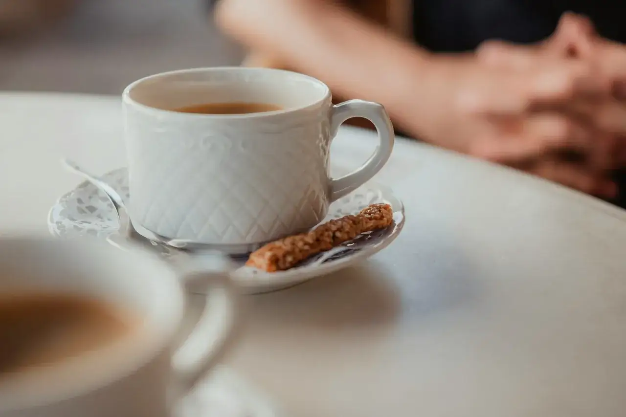 Eine Tasse Mokka mit Milch und ein Keks auf einem Unterteller. Im Hintergrund sind Hände zu sehen.