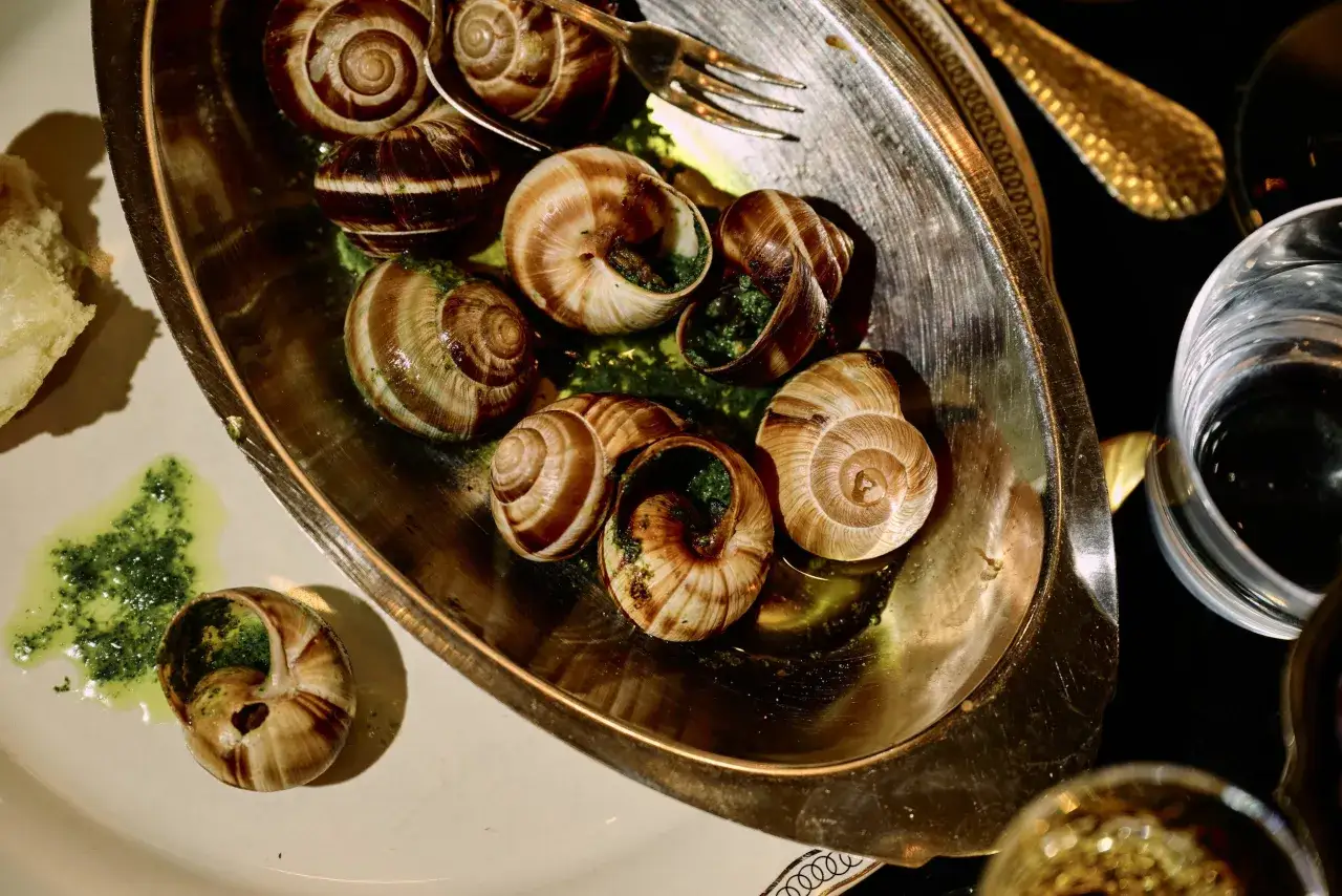 Caracoles con mantequilla y perejil, servidos en una bandeja metálica, listos para ser disfrutados con un tenedor.
