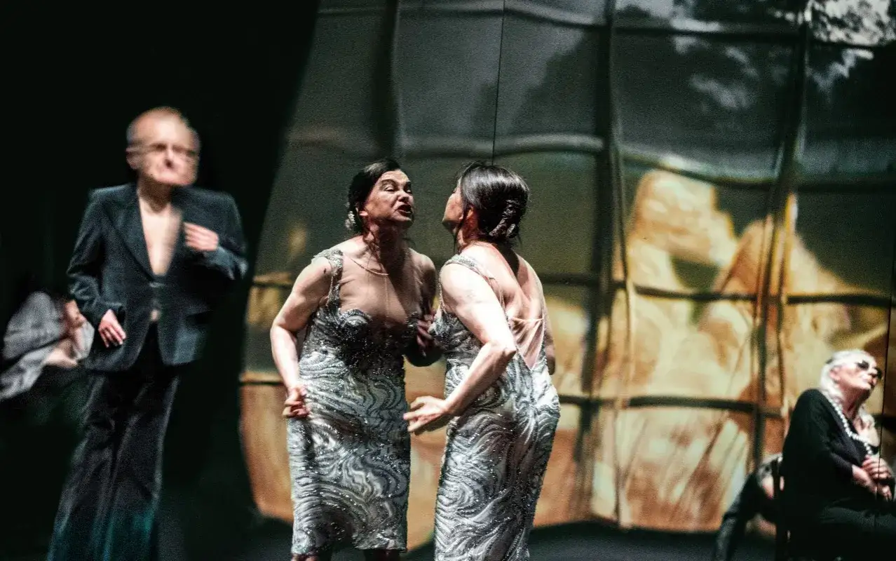 Aktorzy Teatru Nowego w Warszawie w błyszczących sukniach, w tle rozmyta postać mężczyzny w marynarce.